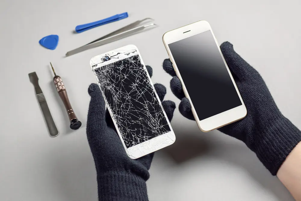 Person hält ein neues und ein beschädigtes Smartphone in den Händen, vor einem Tisch auf dem Instrumente für die Reparatur liegen.